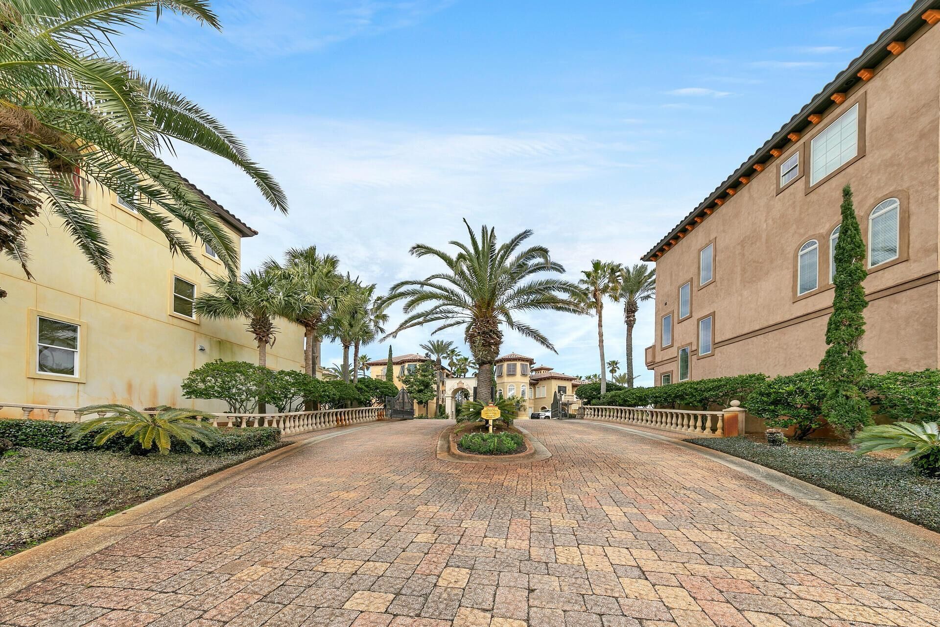 A paved pathway leading to a building with a central circular garden, flanked by palm trees and a stone balustrade.