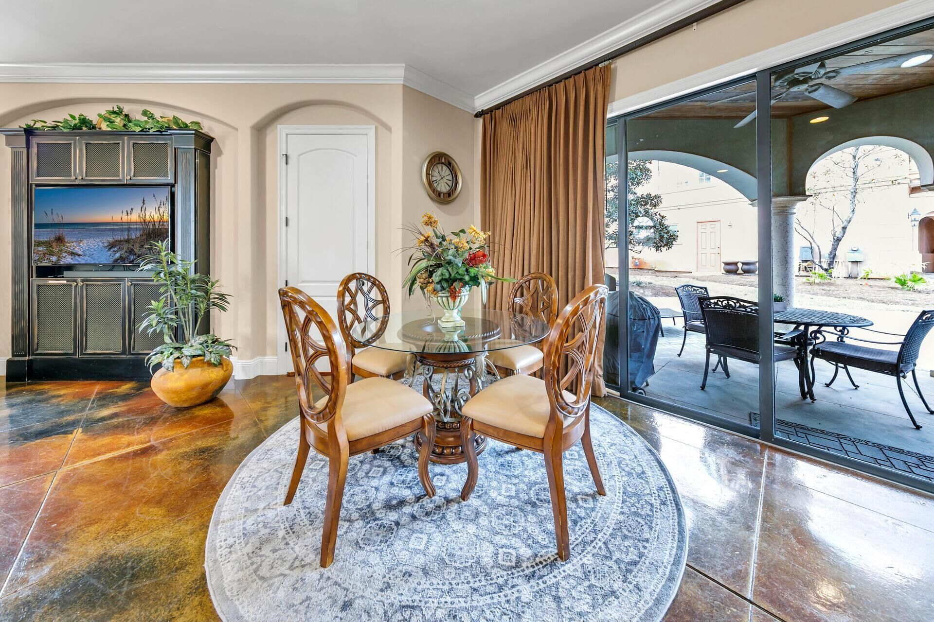 A dining area with a round table, four chairs, a vase with flowers, and a large window with curtains.