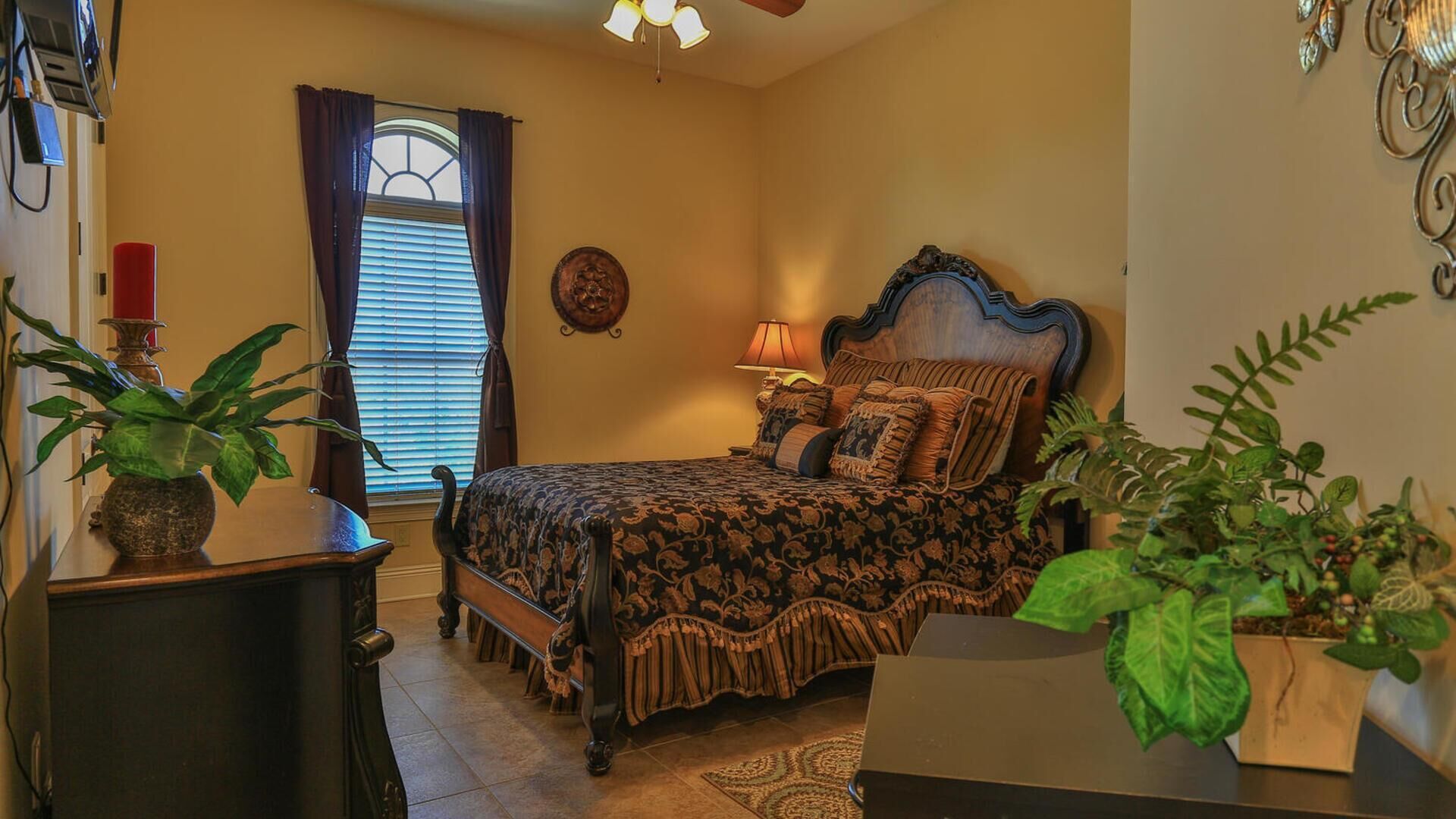 A bedroom with a bed, a nightstand, a lamp, a window with curtains, and two potted plants.