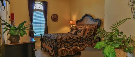 A bedroom with a bed, a nightstand, a lamp, a window with curtains, and two potted plants.