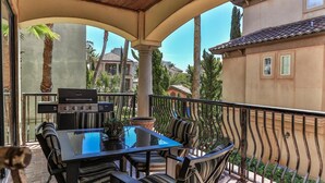 A covered outdoor dining area with a grill, striped cushions, and a view of the surrounding buildings and palm trees. - Tuscany (Miramar Beach)