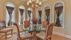 A dining area with a glass table surrounded by four chairs, a chandelier, and large windows with curtains. - Tuscany (Miramar Beach)
