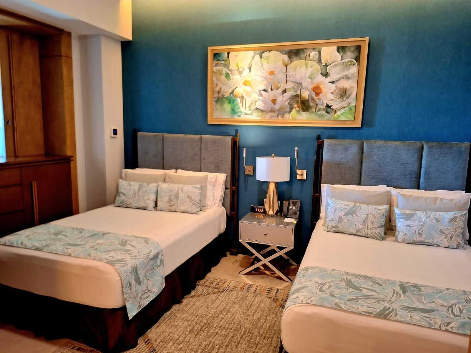 Ett hotellrum med två sängar, ett nattduksbord med en lampa, en tavla med blommor på väggen och en garderob i trä.