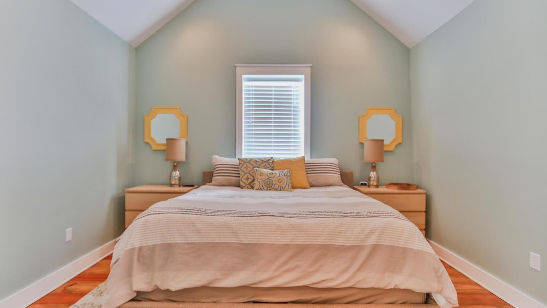 A bedroom with a sloped ceiling, a large bed with a beige bedspread, two nightstands with lamps, and a window with blinds.