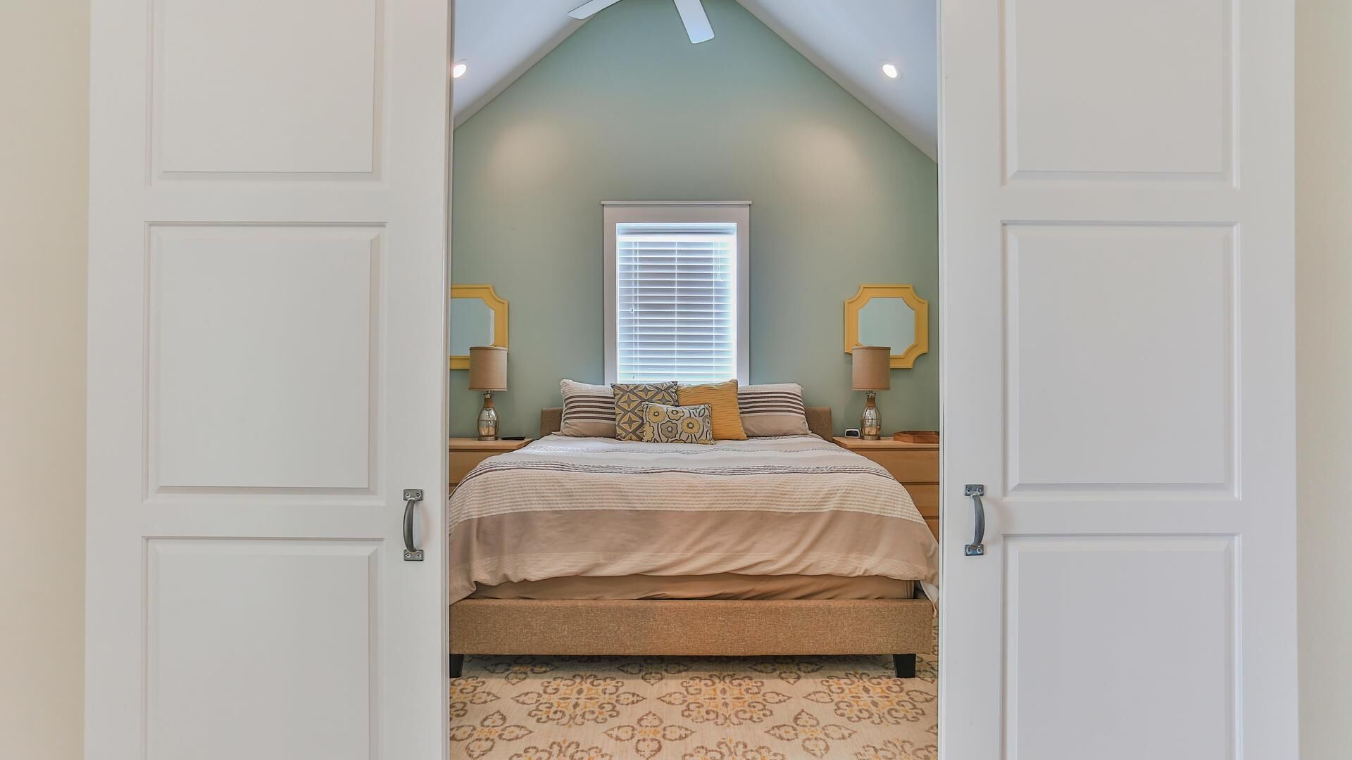 A bedroom with a sloped ceiling, a bed with patterned bedding, two bedside tables with lamps, and a window with blinds.