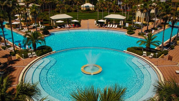 Une grande piscine circulaire avec une fontaine centrale, entourée de palmiers et de chaises longues.