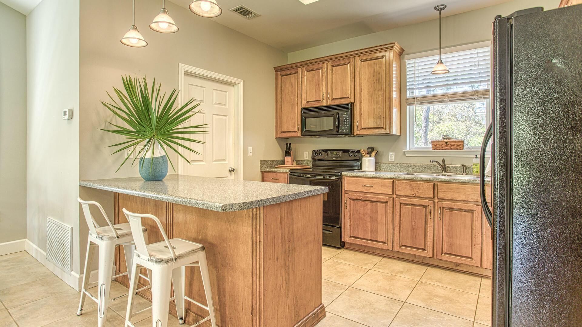 A kitchen with wooden cabinets, a black refrigerator, a microwave, and a sink. There is a potted plant on the counter and two white bar stools.