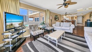 A spacious living room with a large TV, comfortable seating, a ceiling fan, and a view of the balcony. - Destin Towers 102 (Destin)