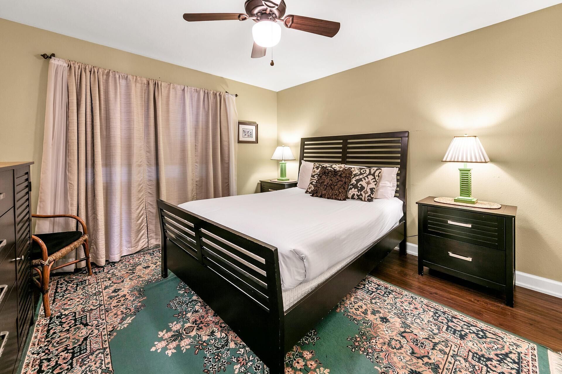 Une chambre à coucher avec un lit, deux tables de chevet avec des lampes, un ventilateur de plafond, une chaise, un bureau et un tapis à motifs.