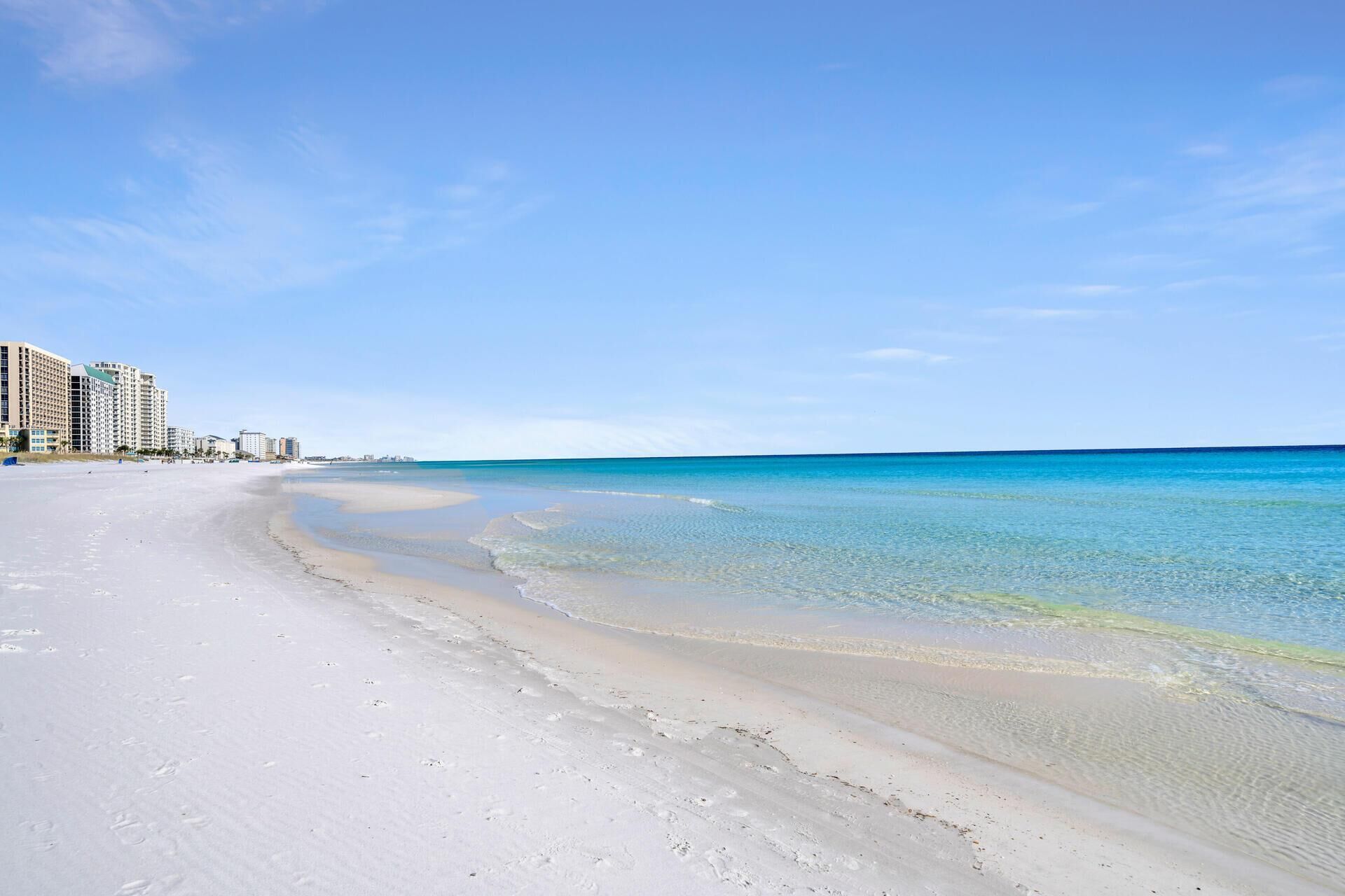 A beach with clear blue water and a sandy shore. There are buildings along the coastline.