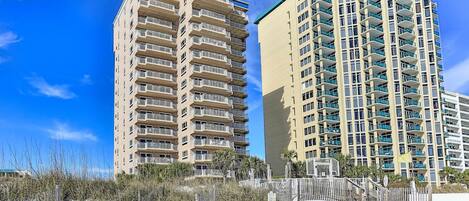 Two high-rise buildings with multiple balconies, a sandy beach in the foreground, and a clear blue sky.