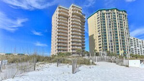 Two high-rise buildings with multiple balconies, a sandy foreground with a fence, and a clear blue sky. - Destin Towers 111 (Destin)