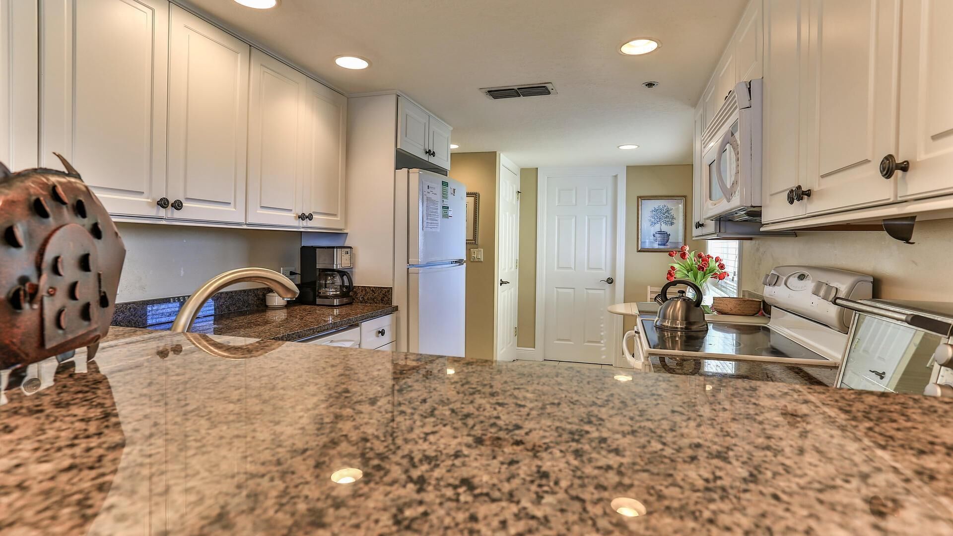 A kitchen with granite countertops, white cabinets, a refrigerator, a coffee maker, and a kettle.