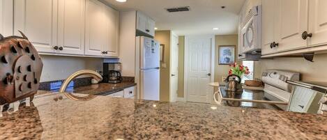 A kitchen with granite countertops, white cabinets, a refrigerator, a coffee maker, and a kettle.
