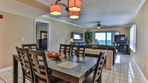 A dining area with a wooden table and chairs, a ceiling fan, and a view of the ocean through large windows. - Destin Towers 142 (Destin)