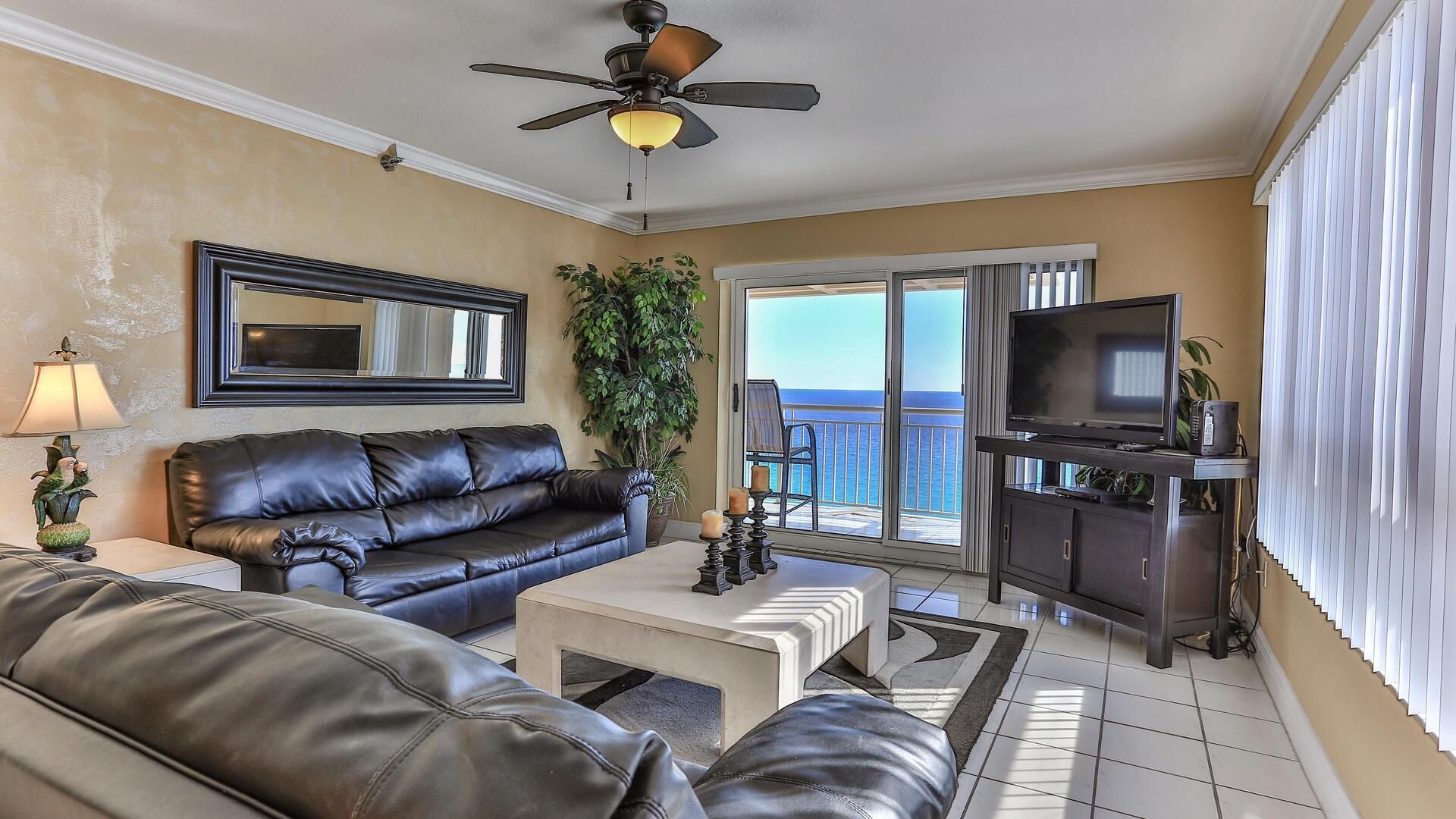 A living room with a black leather sofa, a coffee table, a TV on a stand, a ceiling fan, and a balcony with a view of the ocean.