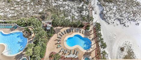 Vista aérea de um resort com duas piscinas, uma área de praia e um espaço de descanso com espreguiçadeiras.