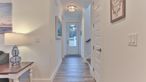 A hallway with a wooden floor, white walls, a door at the end, and a lamp on the left.