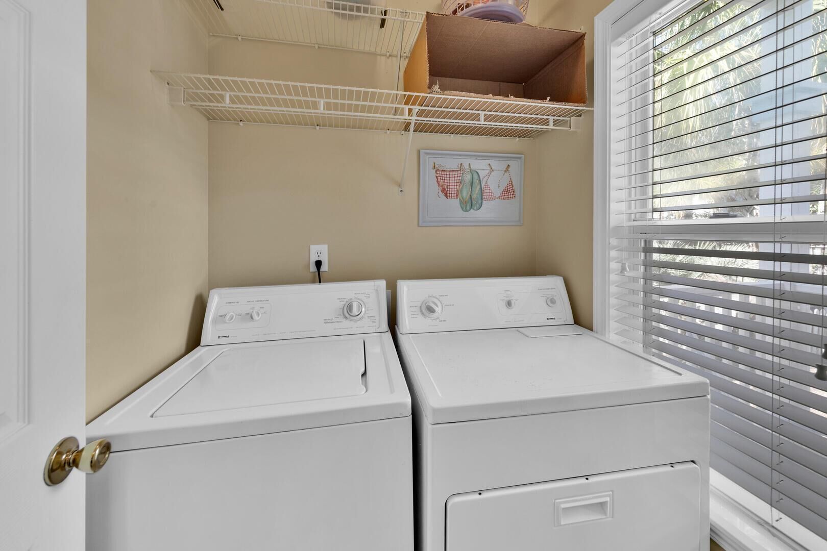 A laundry room with two front-loading washers and dryers, a window with blinds, and a shelf above the dryers.
