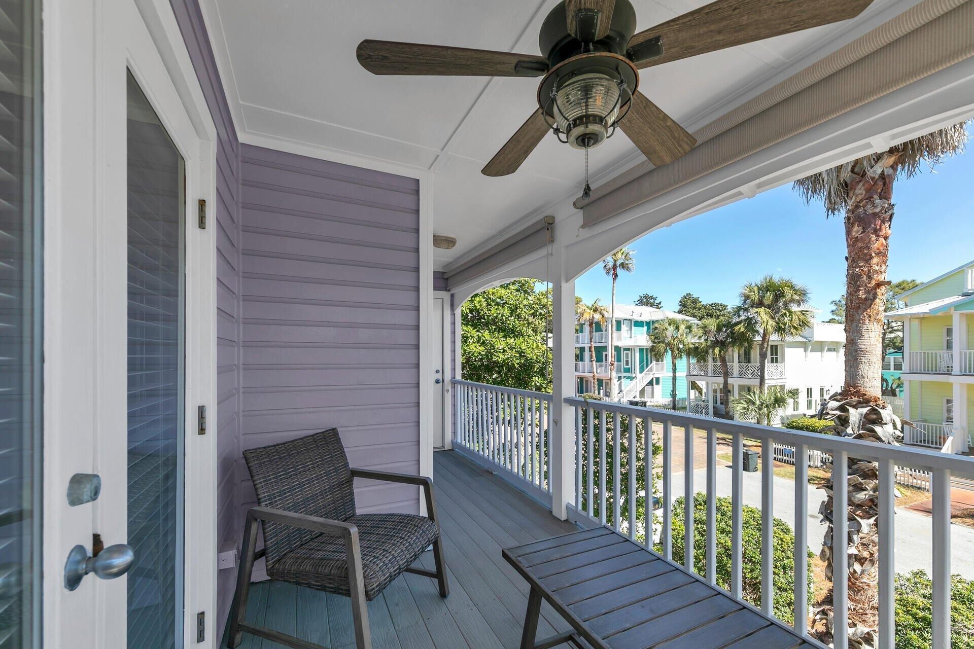 A porch with a ceiling fan, a chair, and a view of a resort area with buildings and palm trees.