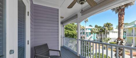 A porch with a ceiling fan, a chair, and a view of a resort area with buildings and palm trees.