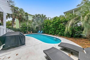 A swimming pool with lounge chairs and a barbecue area, surrounded by lush greenery and a building in the background. - La Bella Vita (Santa Rosa Beach)