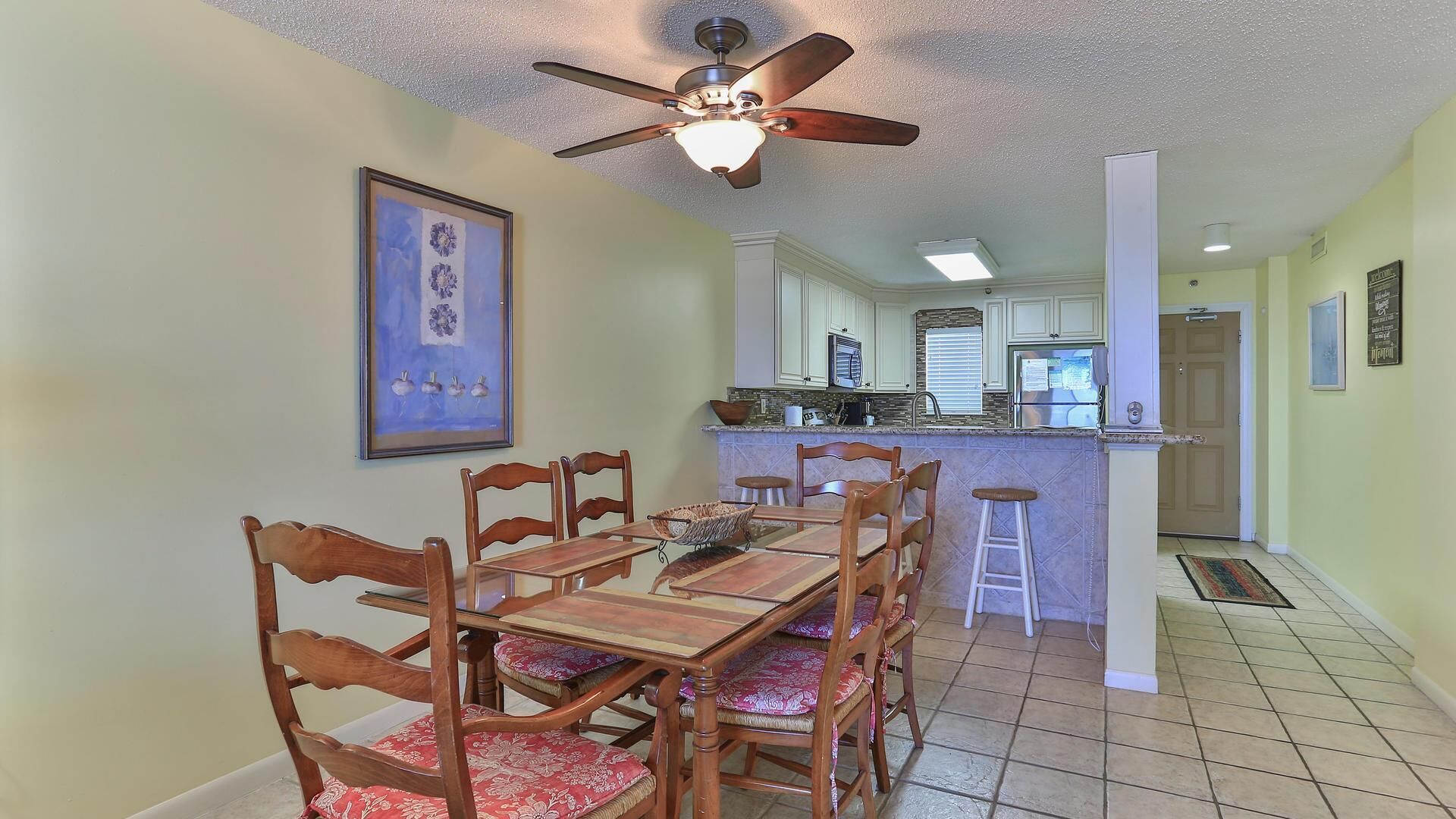 A dining area with a wooden table and chairs, a ceiling fan, and a kitchen in the background.