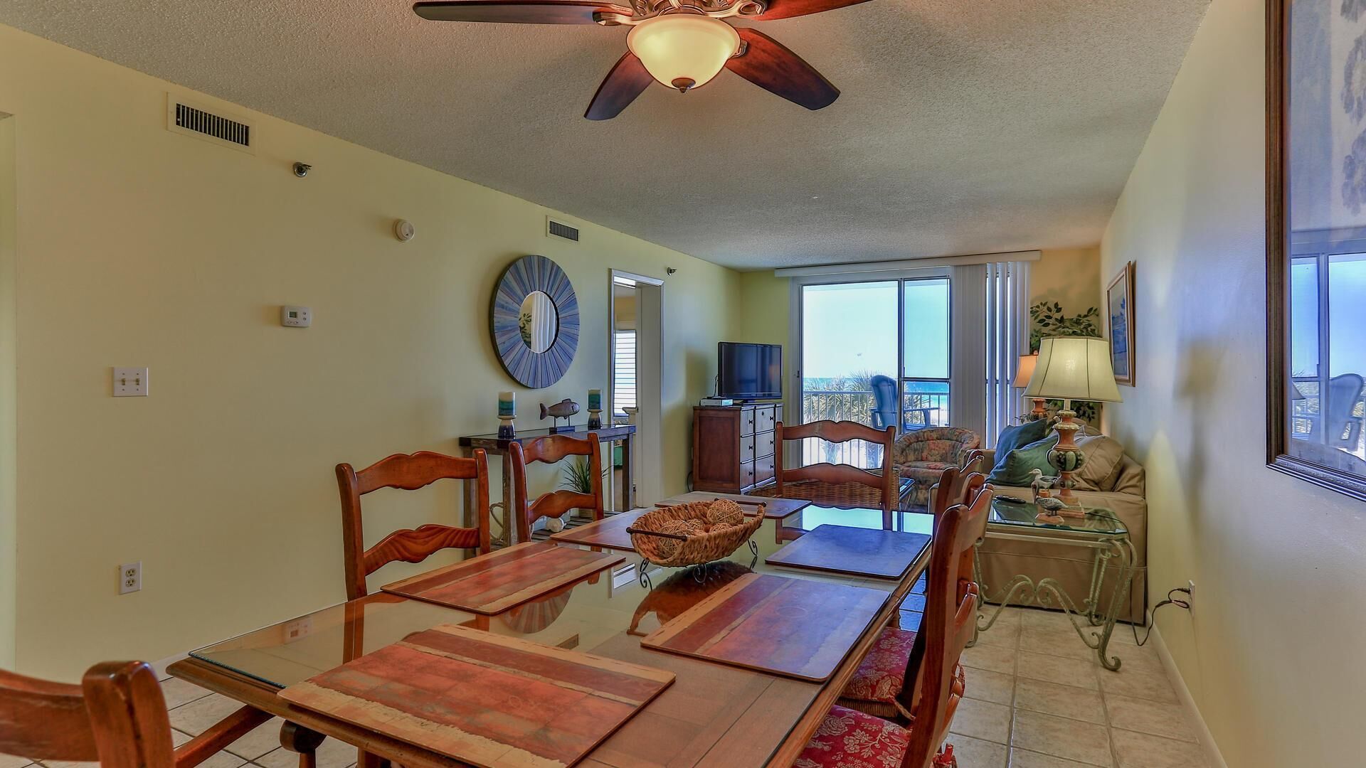 A dining area with a wooden table and chairs, a ceiling fan, and a view of the outdoors through large windows.