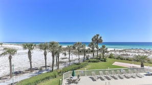 A beach view with palm trees, a fenced patio area with lounge chairs, and a clear blue sky. - Destin on the Gulf 302 (Destin)