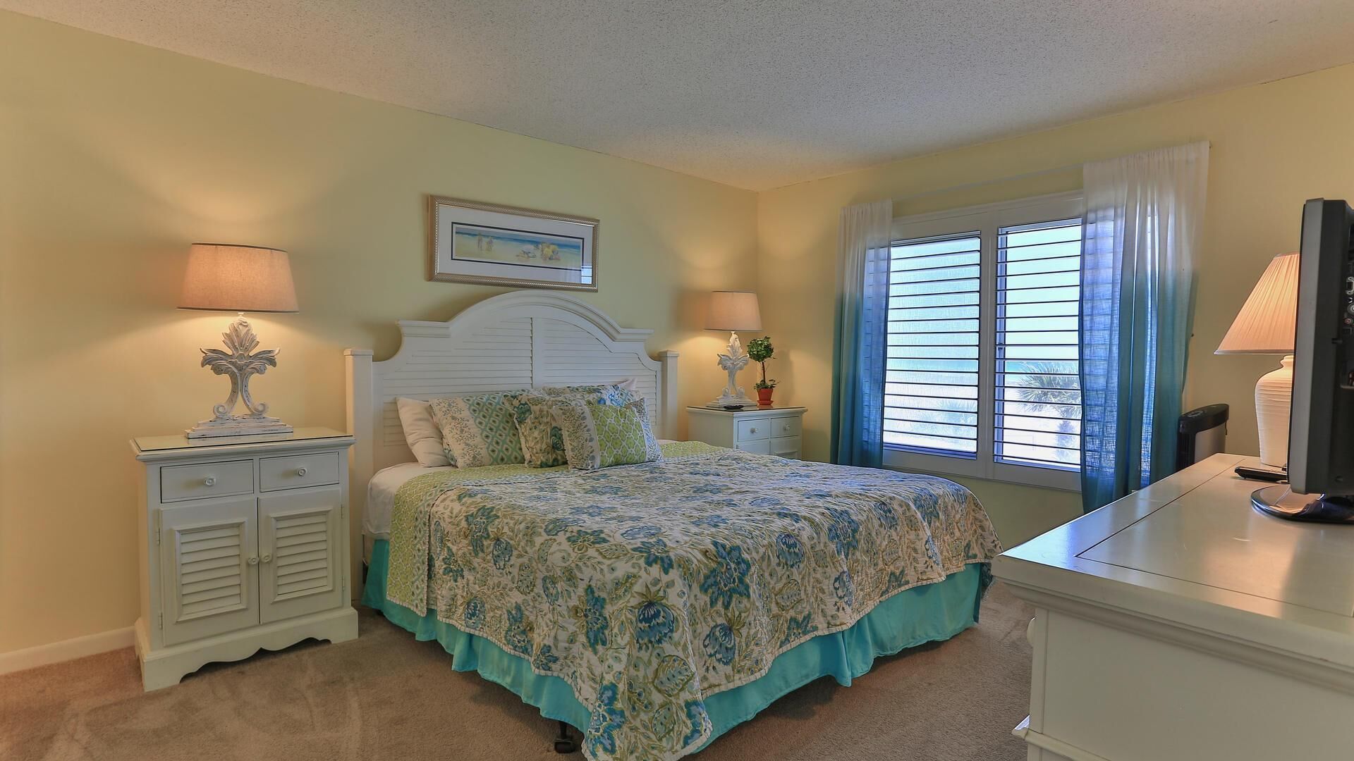 A bedroom with a bed, two bedside tables, a TV, and a window with blinds.