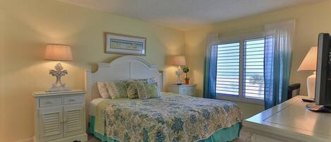 A bedroom with a bed, two bedside tables, a TV, and a window with blinds.