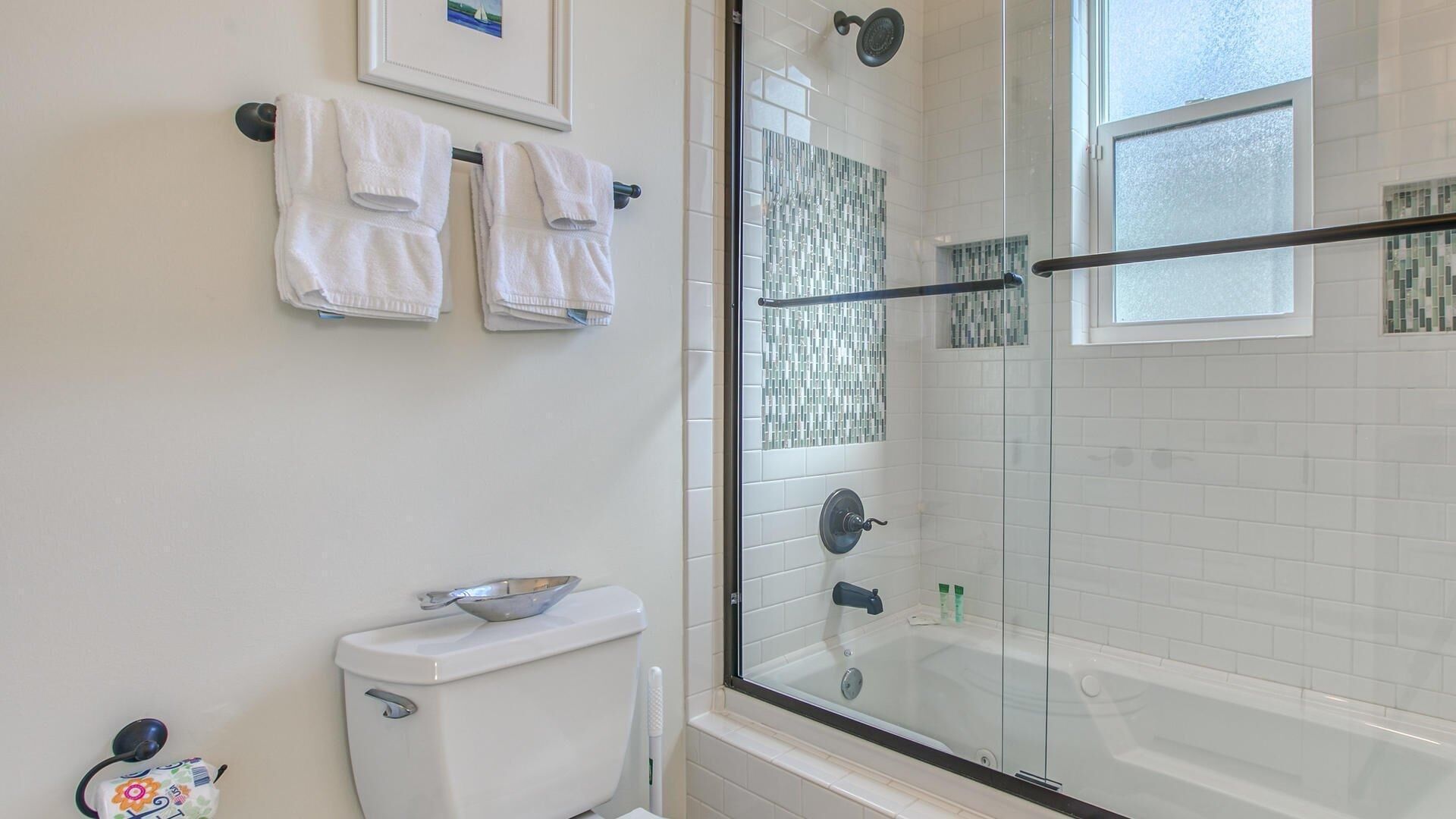 Un baño con ducha compartimentada con vidrio, azulejos blancos, un inodoro y un cuadro en la pared.