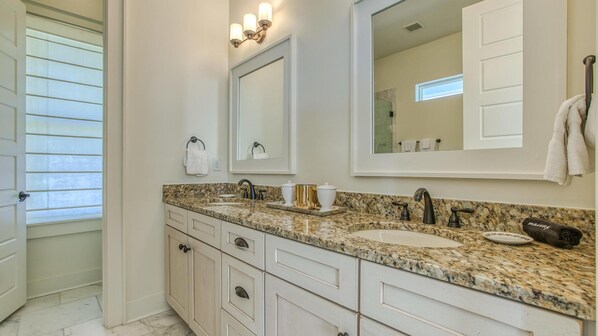 A bathroom with a granite countertop, white cabinetry, a large mirror, and a window with blinds.