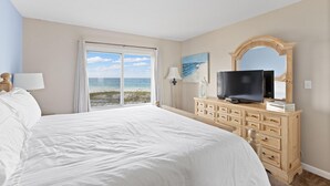 Een slaapkamer met een groot bed, een televisie op een houten dressoir, een schilderij van een strand en uitzicht op de oceaan door een raam.
