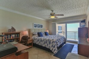 A hotel room with a bed, a desk with a laptop, a bookshelf, a ceiling fan, and a view of the ocean through a balcony.