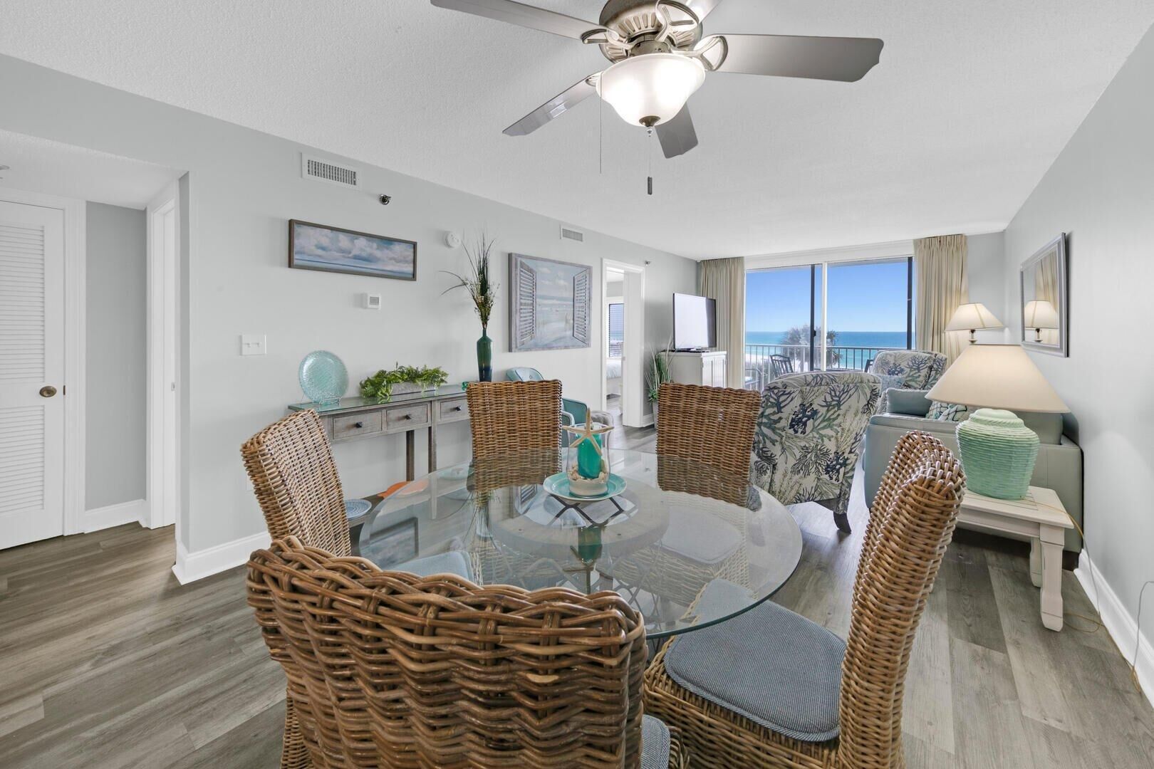 A modern living room with a glass dining table surrounded by wicker chairs, a ceiling fan, and a view of the ocean.
