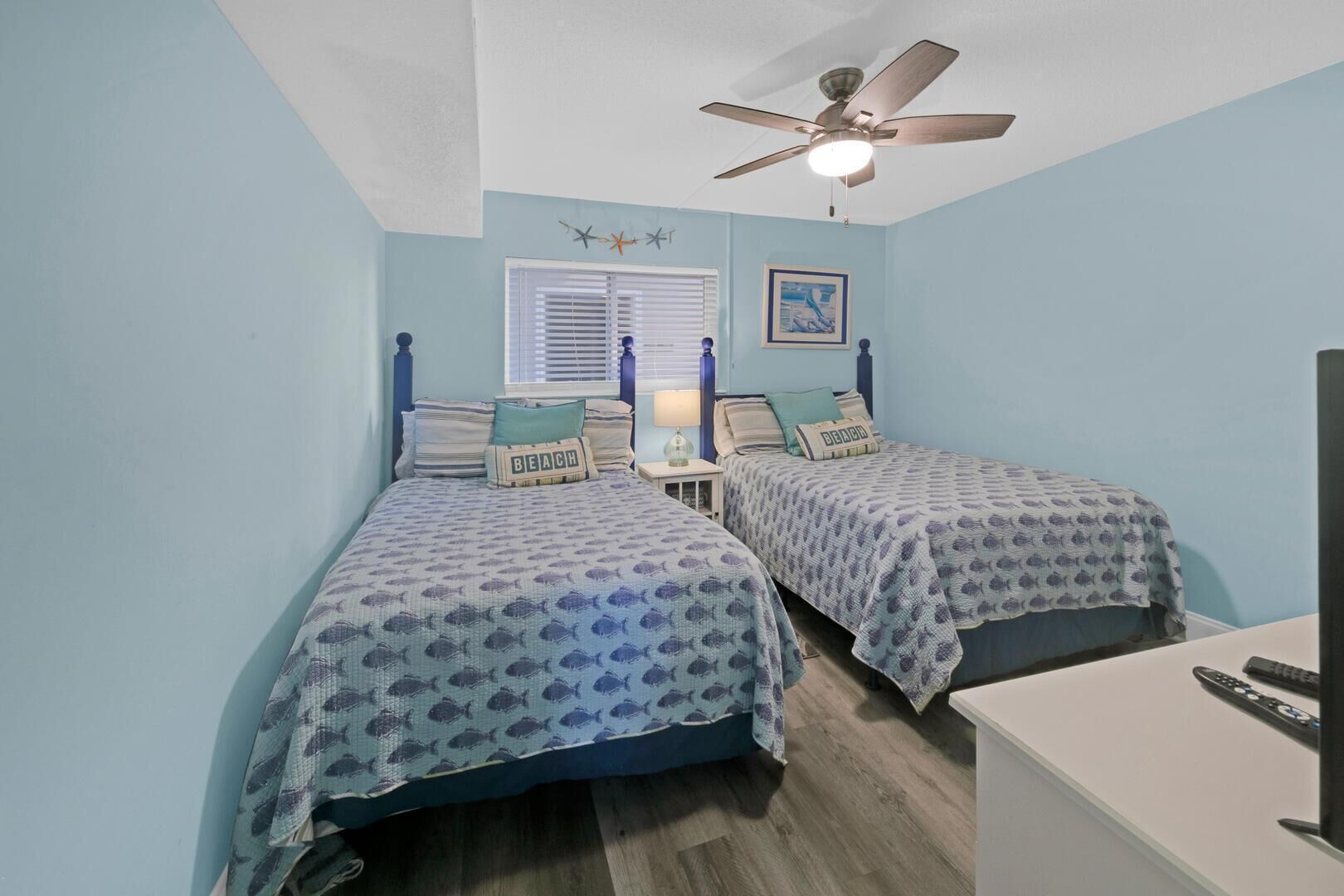 A bedroom with two beds, a ceiling fan, a window with blinds, and a framed picture on the wall.