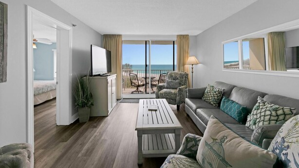 Un salon moderne avec un canapé, une table basse et des fauteuils, un balcon offrant une vue sur la plage et un téléviseur fixé au mur.