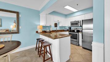 A kitchen with white cabinets, a granite countertop, a stainless steel refrigerator, and a dining area with a table and bar stools.