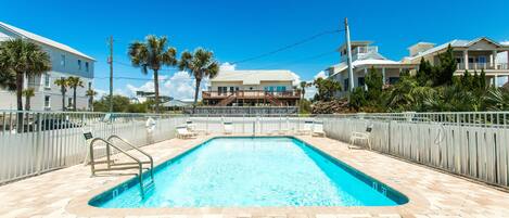 A rectangular swimming pool with a tiled deck, surrounded by a metal fence, and palm trees in the background.