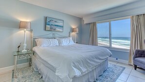 A large bed with white linens, a painting of a beach scene, and a view of the ocean through a large window. - Destin on the Gulf 703 (Destin)