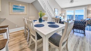 Un coin repas avec une table et des chaises blanches, un vase de fleurs et une vue sur l’océan par de grandes fenêtres.