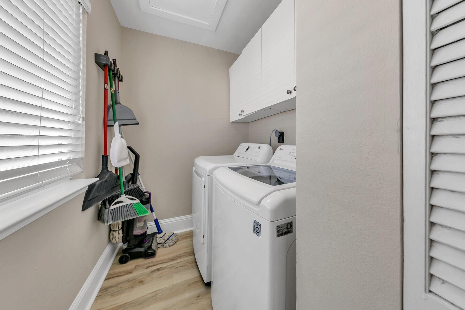 A laundry room with a washing machine and dryer, a mop and broom, and a window with blinds.