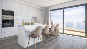 A modern kitchen with white cabinetry, a built-in oven, and a dining area with a large window overlooking a scenic view. - Sunshine Deluxe B by Madeira Sun Travel (Ponta do Sol)