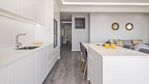 A modern kitchen with white cabinets, a central island, and a dining area with a grey sofa. - Sunshine Deluxe C by Madeira Sun Travel (Ponta do Sol)