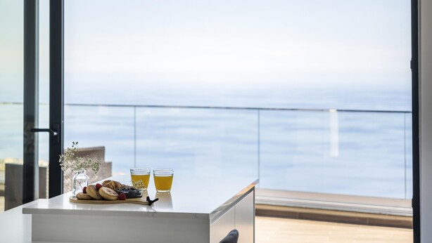 A breakfast table with pastries, a glass of juice, and a vase with flowers, overlooking a balcony with a view of the sea. - Sunshine Deluxe C by Madeira Sun Travel (Ponta do Sol)