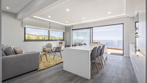A modern living room with a large island table, grey sofa, and a view of the sea. - Sunshine Deluxe C by Madeira Sun Travel (Ponta do Sol)