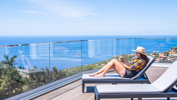 A person relaxing on a sun lounger with a view of the ocean and a distant town. - Sunshine Deluxe D by Madeira Sun Travel (Ponta do Sol)