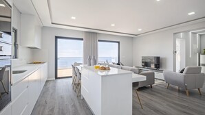 A modern kitchen with a central island, a dining area with a sofa, and a large window offering a view of the sea. - Sunshine Deluxe D by Madeira Sun Travel (Ponta do Sol)