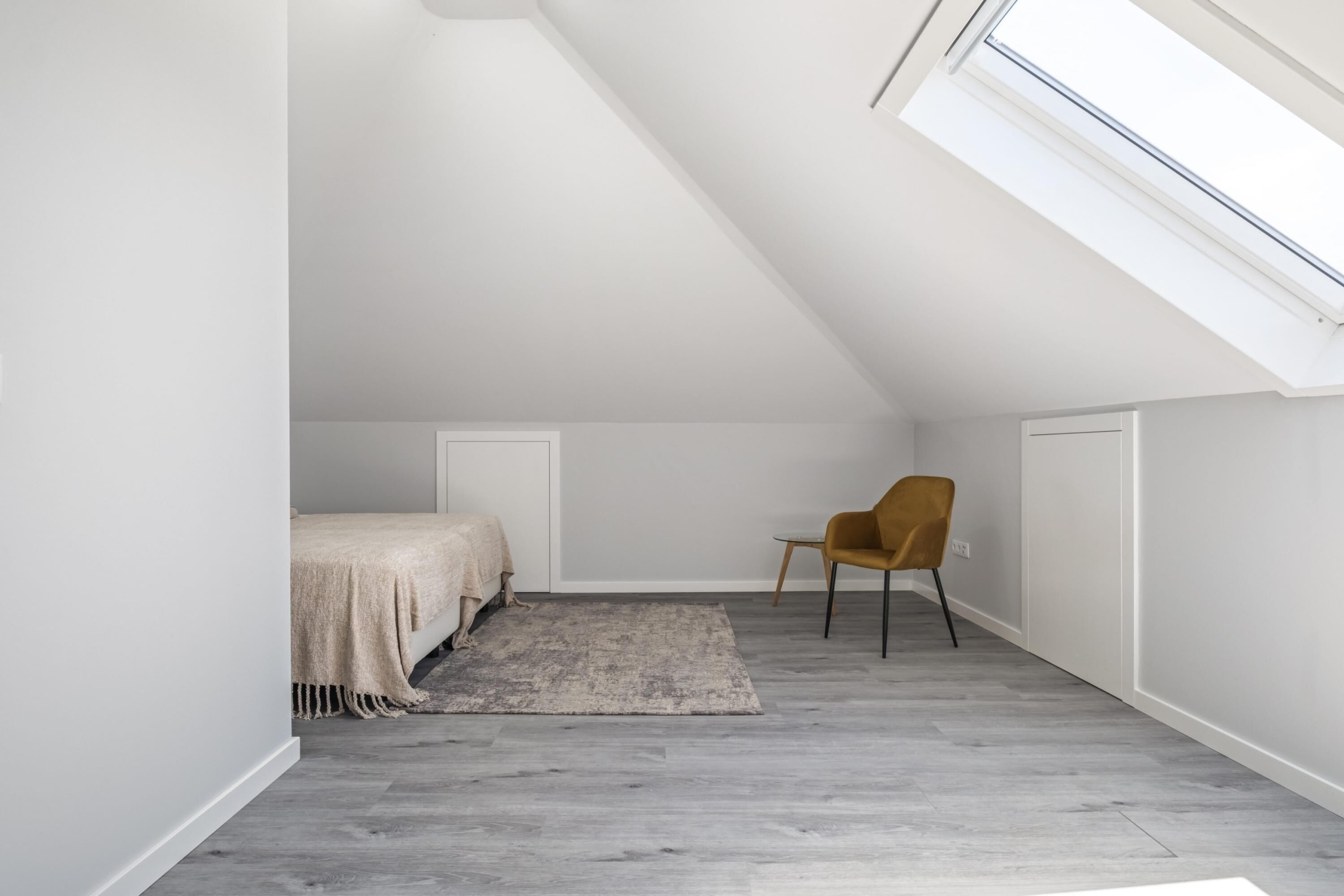 Ein kleines, modernes Schlafzimmer mit schrägem Dach, einem Bett, einem Stuhl und einem Oberlicht.